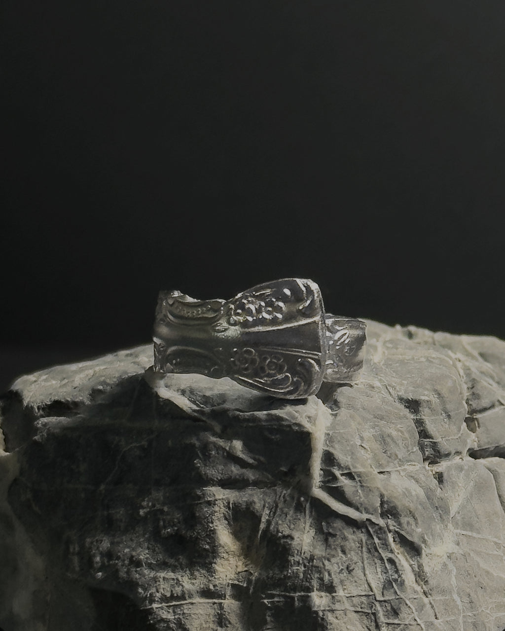 Solid stainless steel spoon ring with ornate engraved floral and scrollwork details, displayed on a textured gray stone against a dark background.