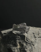 Solid stainless steel spoon ring with ornate engraved floral and scrollwork details, displayed on a textured gray stone against a dark background.