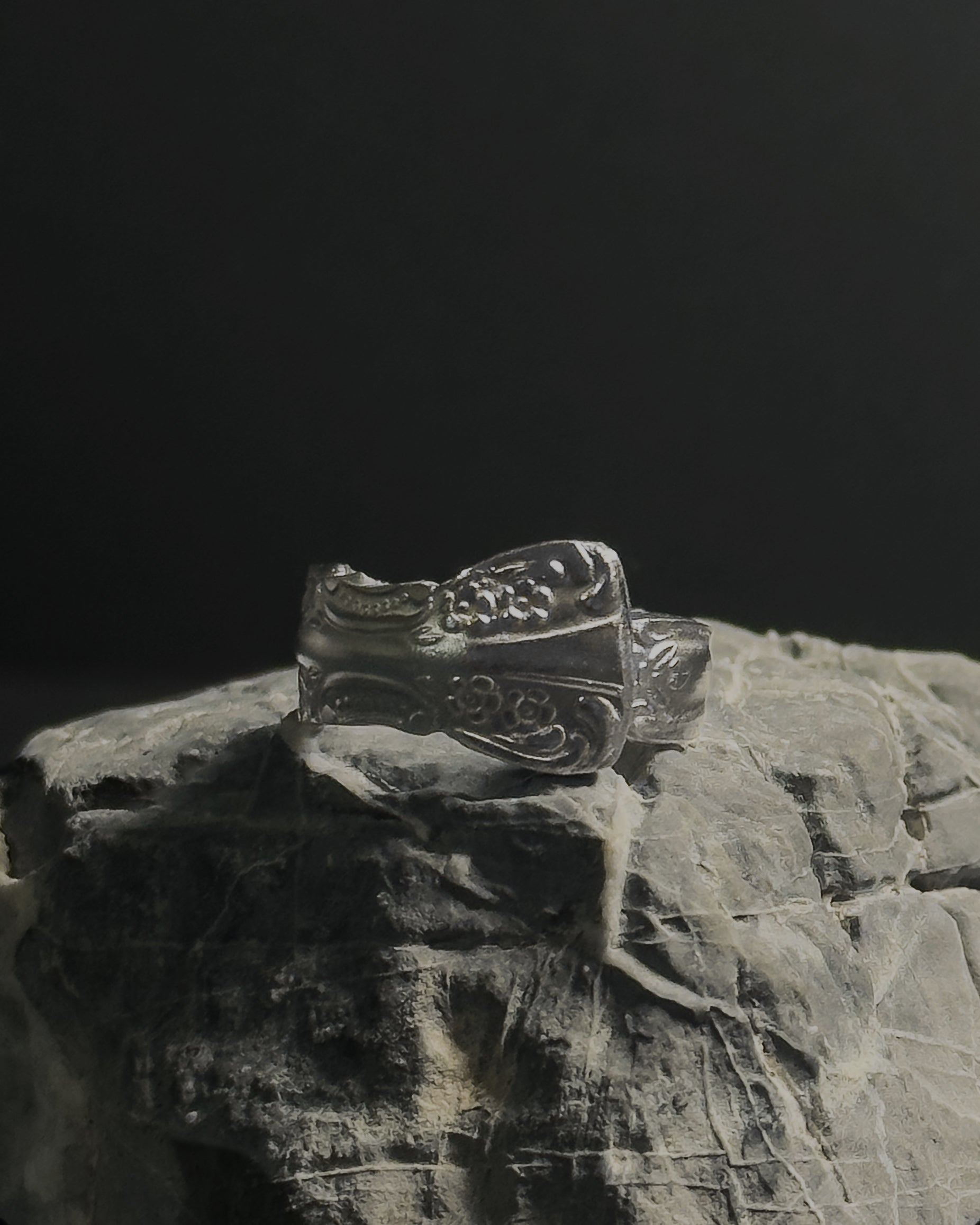 Solid stainless steel spoon ring with ornate engraved floral and scrollwork details, displayed on a textured gray stone against a dark background.