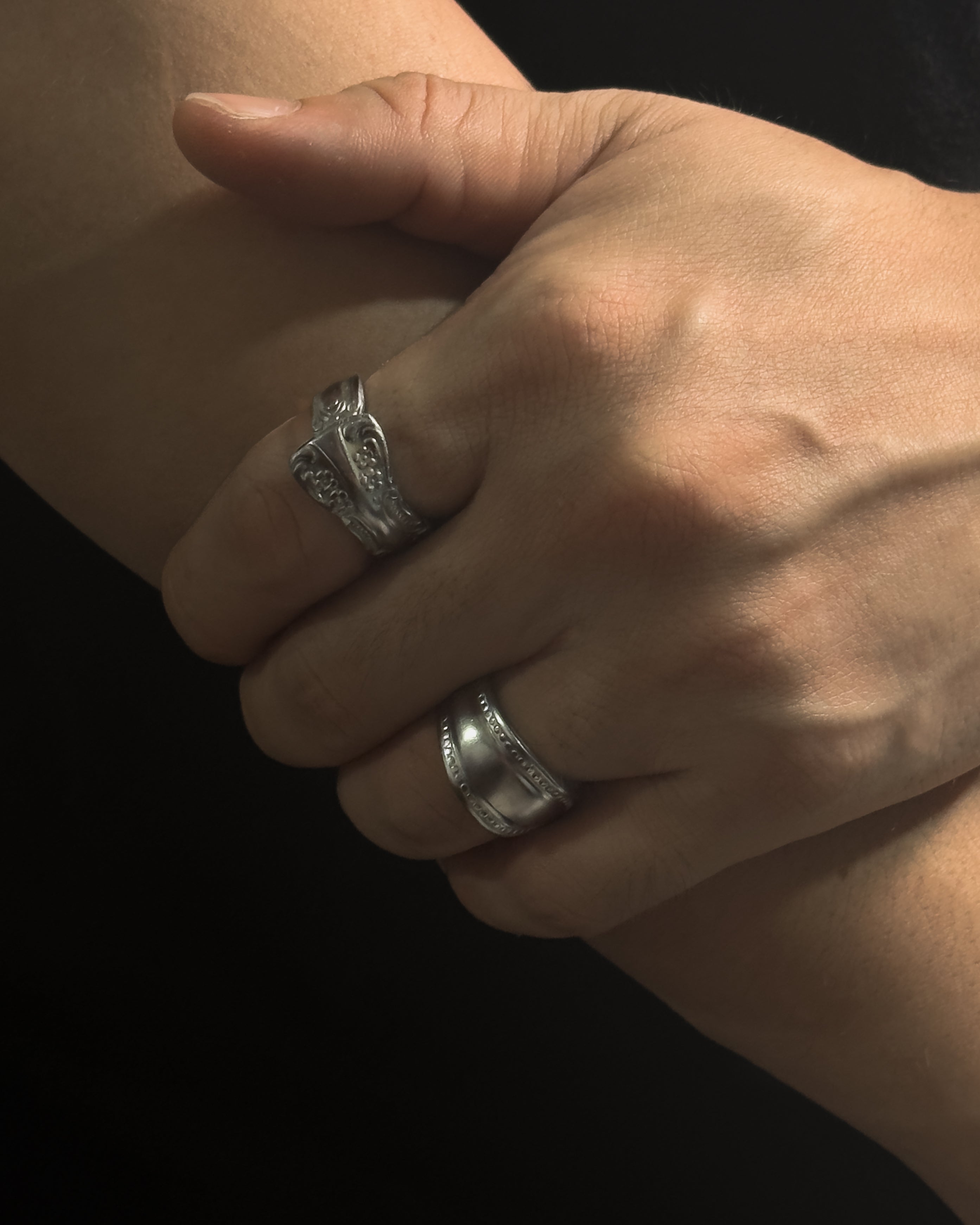 A close-up of a person's hand and forearm wearing two Solid stainless steel spoon ring handcrafted from vintage silverware patterns in a fixed size against a black background.