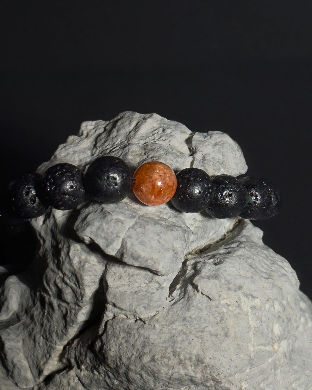 A beaded bracelet made of black lava stone beads with a single round, polished sunstone center bead, displayed on a textured gray rock against a dark background. 
