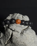 A beaded bracelet made of black lava stone beads with a single round, polished sunstone center bead, displayed on a textured gray rock against a dark background. 