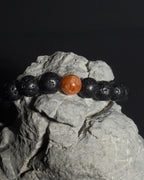 A beaded bracelet made of black lava stone beads with a single round, polished sunstone center bead, displayed on a textured gray rock against a dark background. 