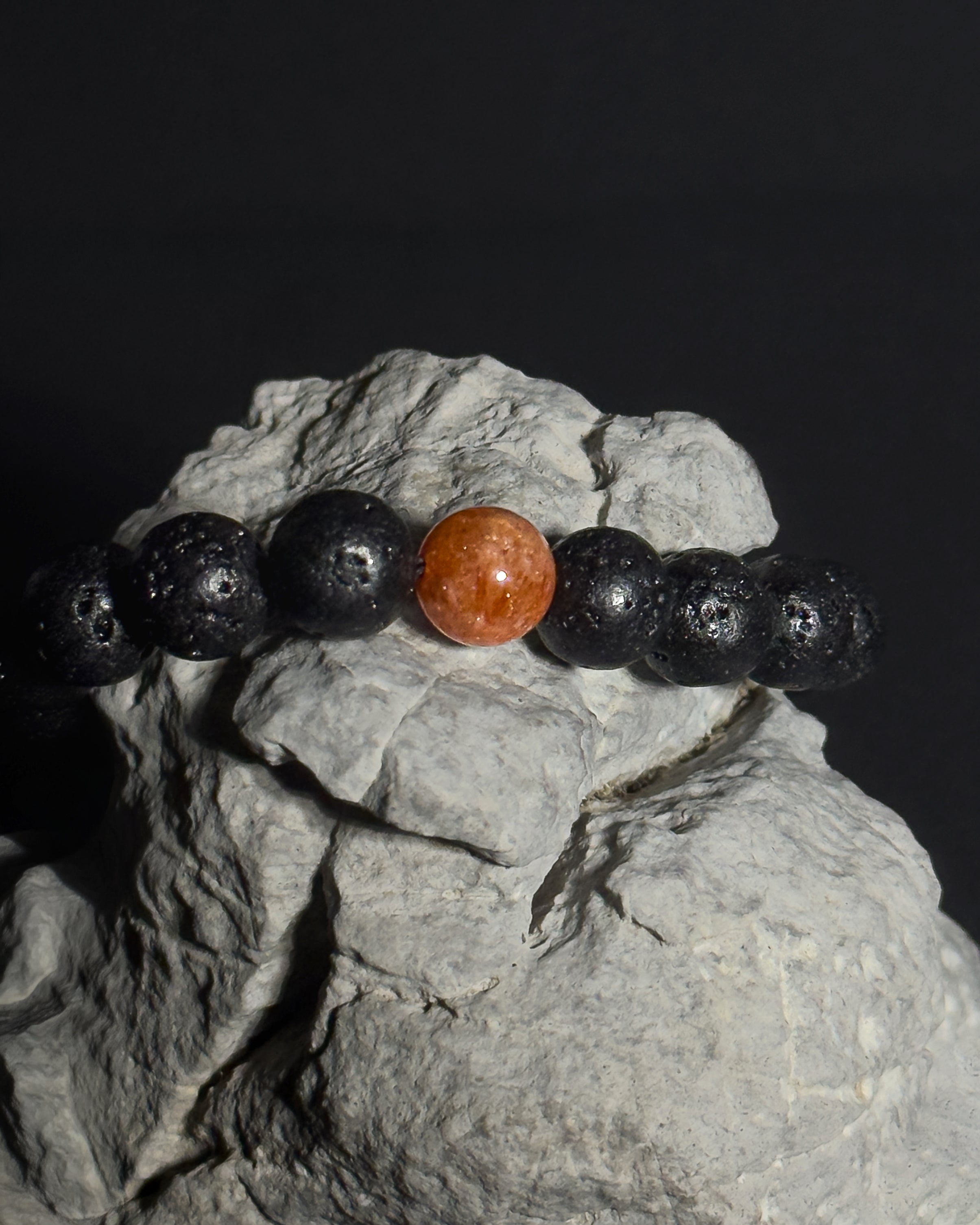 A beaded bracelet made of black lava stone beads with a single round, polished sunstone center bead, displayed on a textured gray rock against a dark background. 