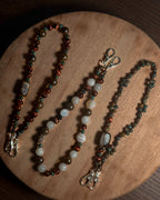 Three beaded phone charm keychain lanyards with silver coloured stainless steel clasp hardware, displayed on a round wooden board. The lanyards feature various stone beads in brown,, bronze, reddish, green, white, and copper tones.