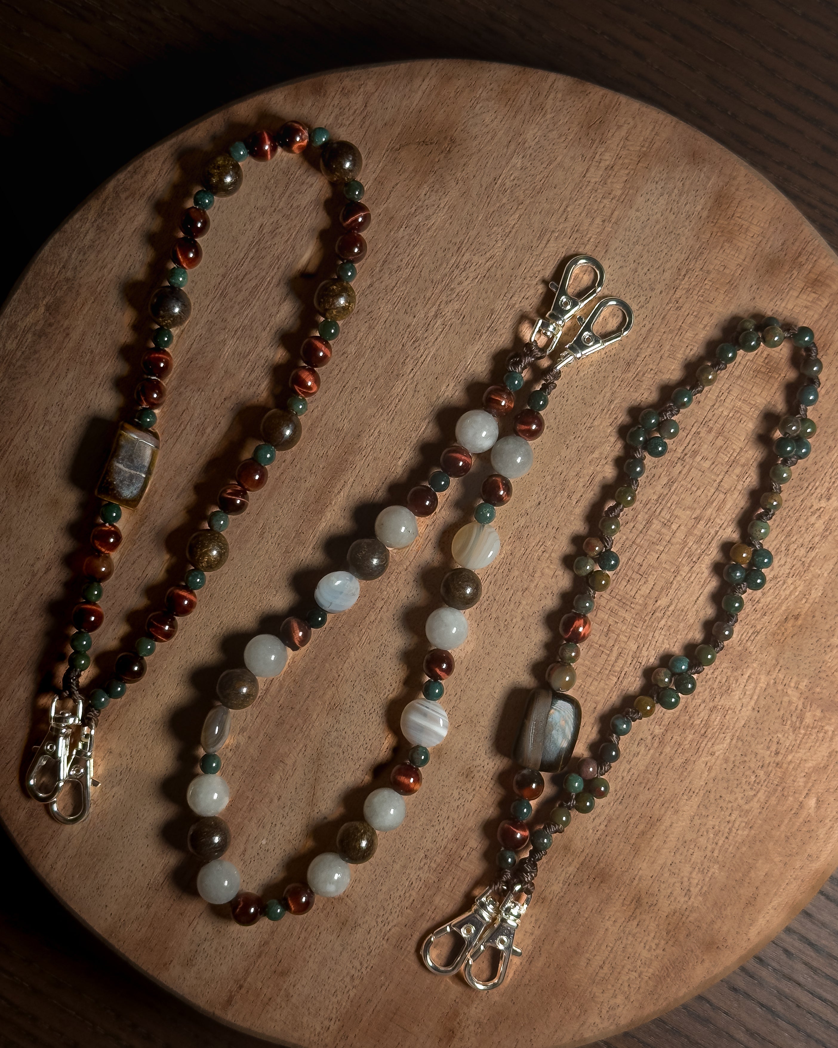 Three beaded phone charm keychain lanyards with silver coloured stainless steel clasp hardware, displayed on a round wooden board. The lanyards feature various stone beads in brown,, bronze, reddish, green, white, and copper tones.