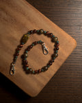 A beaded phone charm with brown, green, and reddish-brown beads, featuring a large polished tiger's eye-like bead and silver-tone stainless steel lobster clasp, displayed on a wooden board.