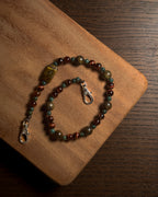 A beaded phone charm with brown, green, and reddish-brown beads, featuring a large polished tiger's eye-like bead and silver-tone stainless steel lobster clasp, displayed on a wooden board.
