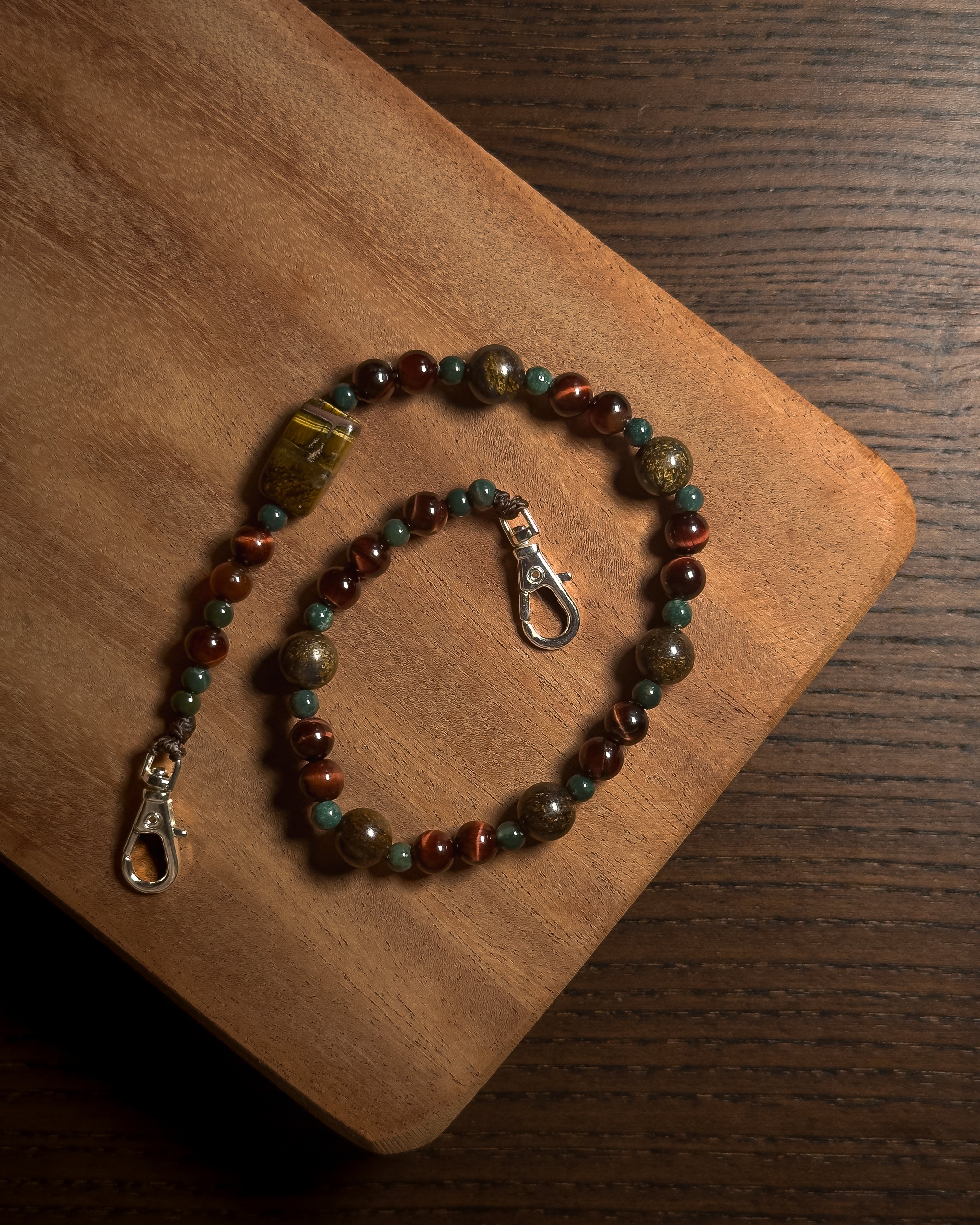 A beaded phone charm with brown, green, and reddish-brown beads, featuring a large polished tiger's eye-like bead and silver-tone stainless steel lobster clasp, displayed on a wooden board.
