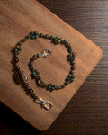 A beaded phone charm with green, brown, and amber beads, featuring a polished tiger's eye accent and silver-tone lobster clasp, displayed on a wooden cutting board.