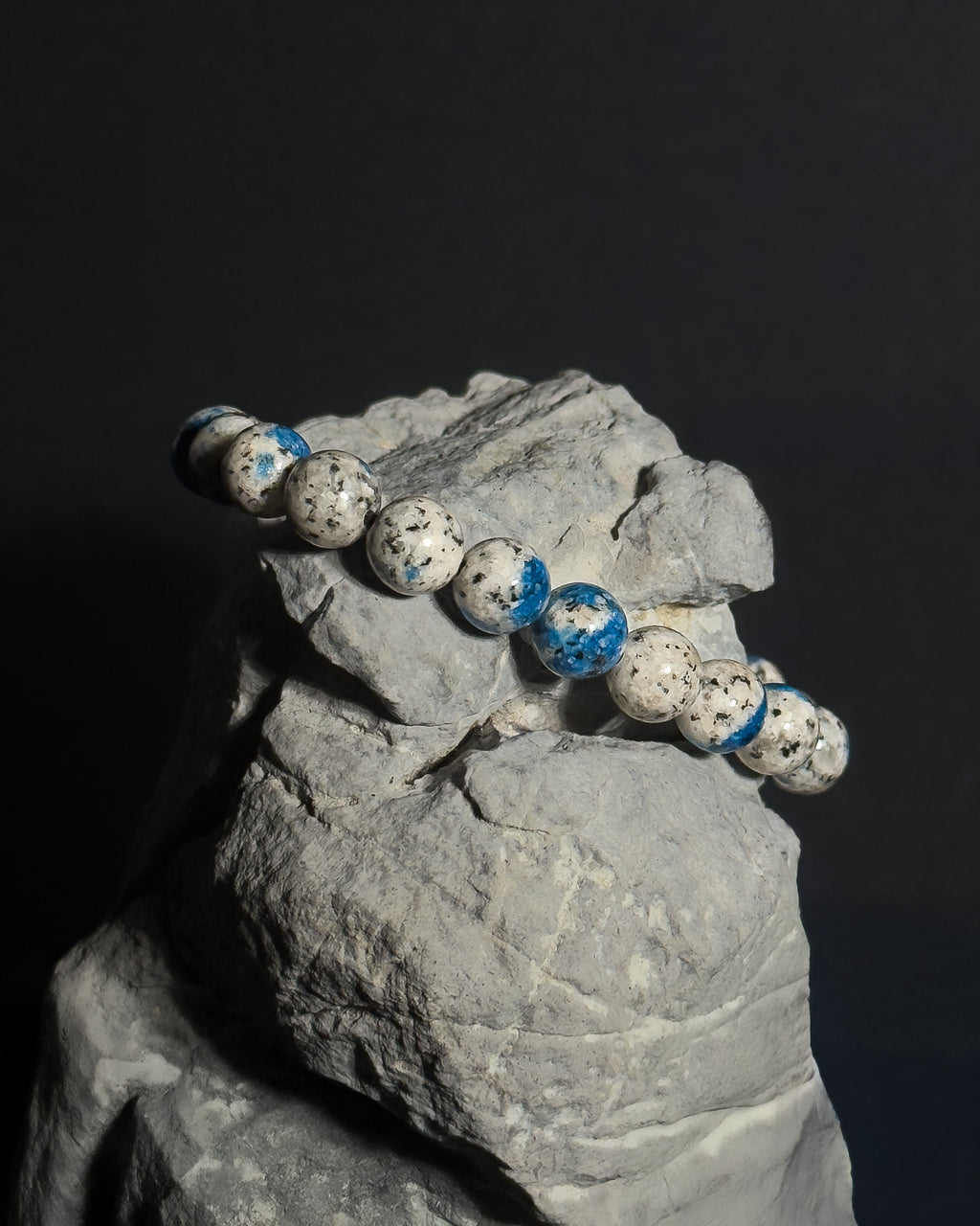A beaded bracelet made of natural K2 stone, draped over a gray rock display against a dark background.
