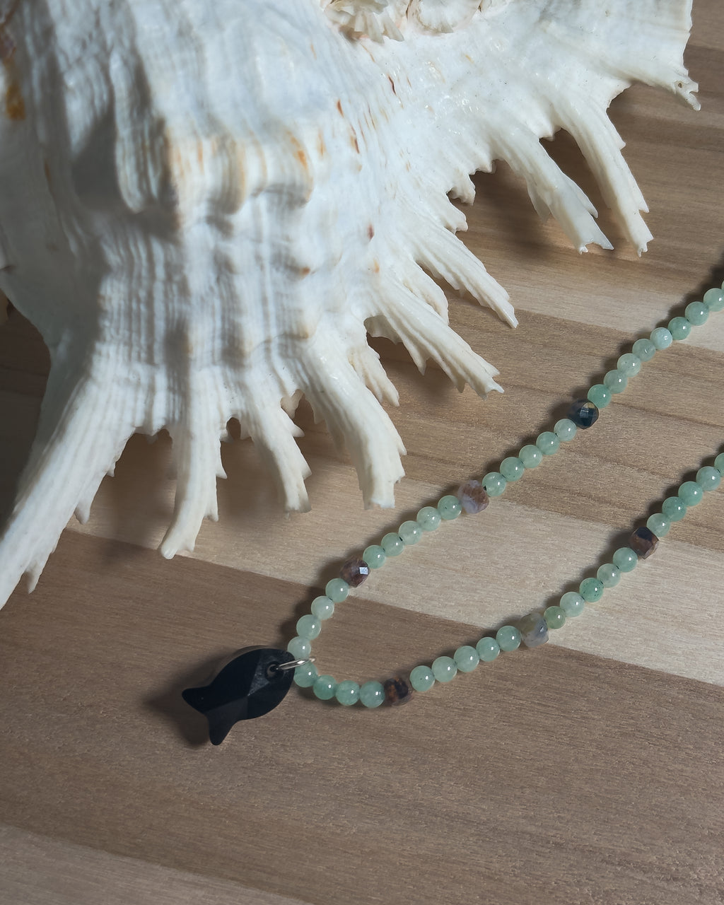 Necklace with a black faceted crystal fish pendant on a wooden surface next to a large shell.