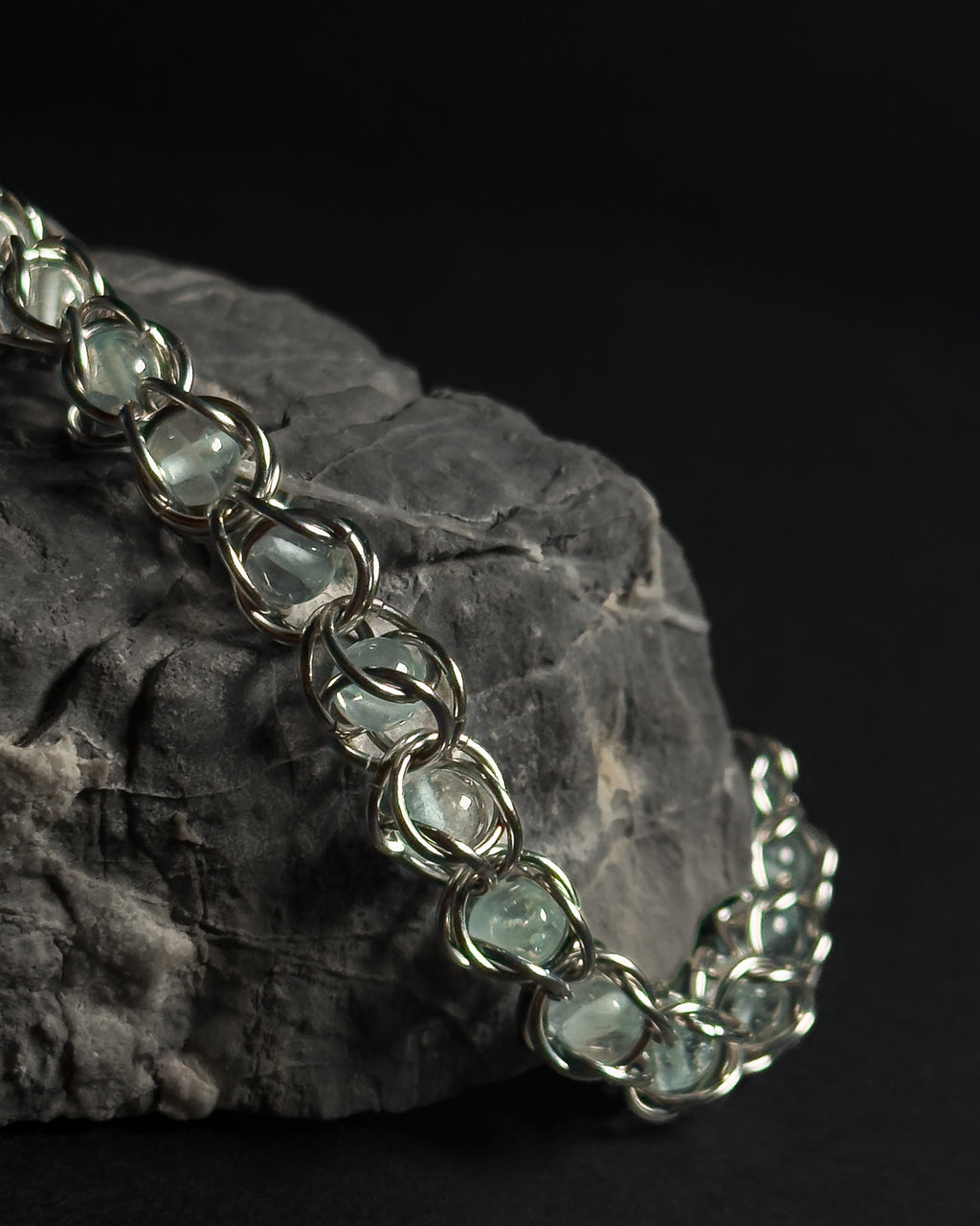 A close-up of a silver-toned chain bracelet with blue topaz captured beads, draped over a dark textured rock against a black background.