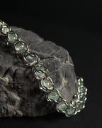 A close-up of a silver-toned chain bracelet with blue topaz captured beads, draped over a dark textured rock against a black background.