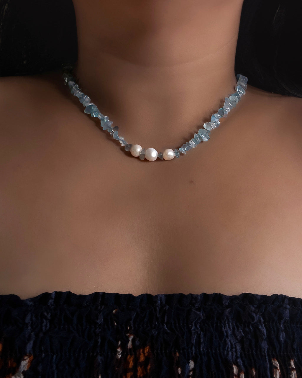 A close-up of a person's neck and chest wearing a beaded choker necklace made of light blue aquamarine faceted chip beads and three white pearl-like beads at the center.