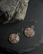 A pair of round silver-toned dangle earrings with colorful, crushed rainbow tourmaline inlays, displayed on a dark slate surface next to a large gray rock.