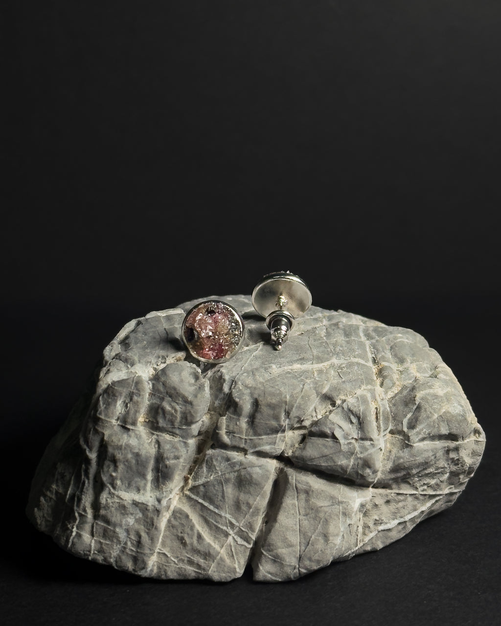 Two round stud earrings with textured, crushed rainbow tourmaline inlay displayed on a gray rock against a solid black background, while the other shows the silver screw-back post.