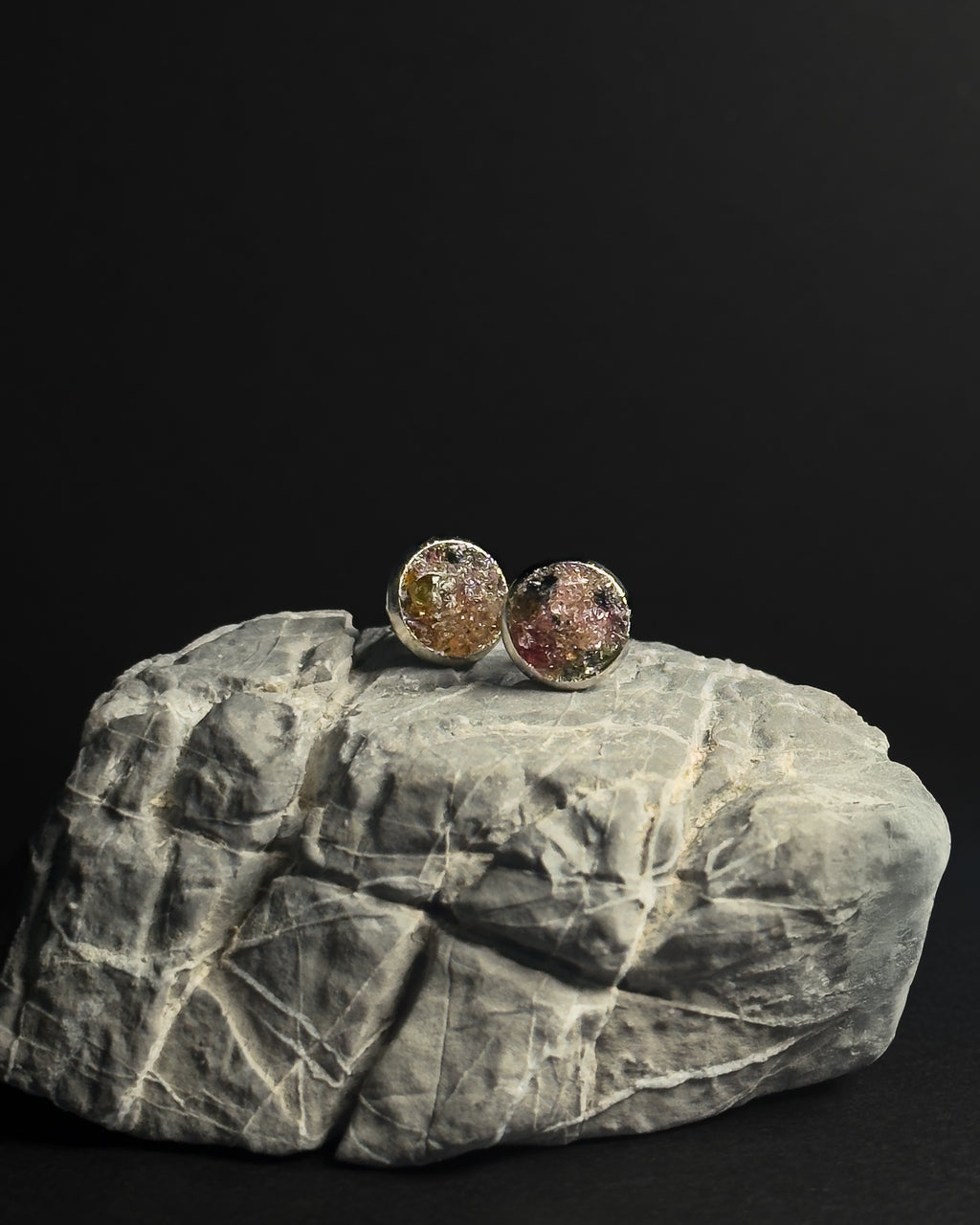 Two round stud earrings with textured, crushed rainbow tourmaline inlay displayed on a gray rock against a solid black background.