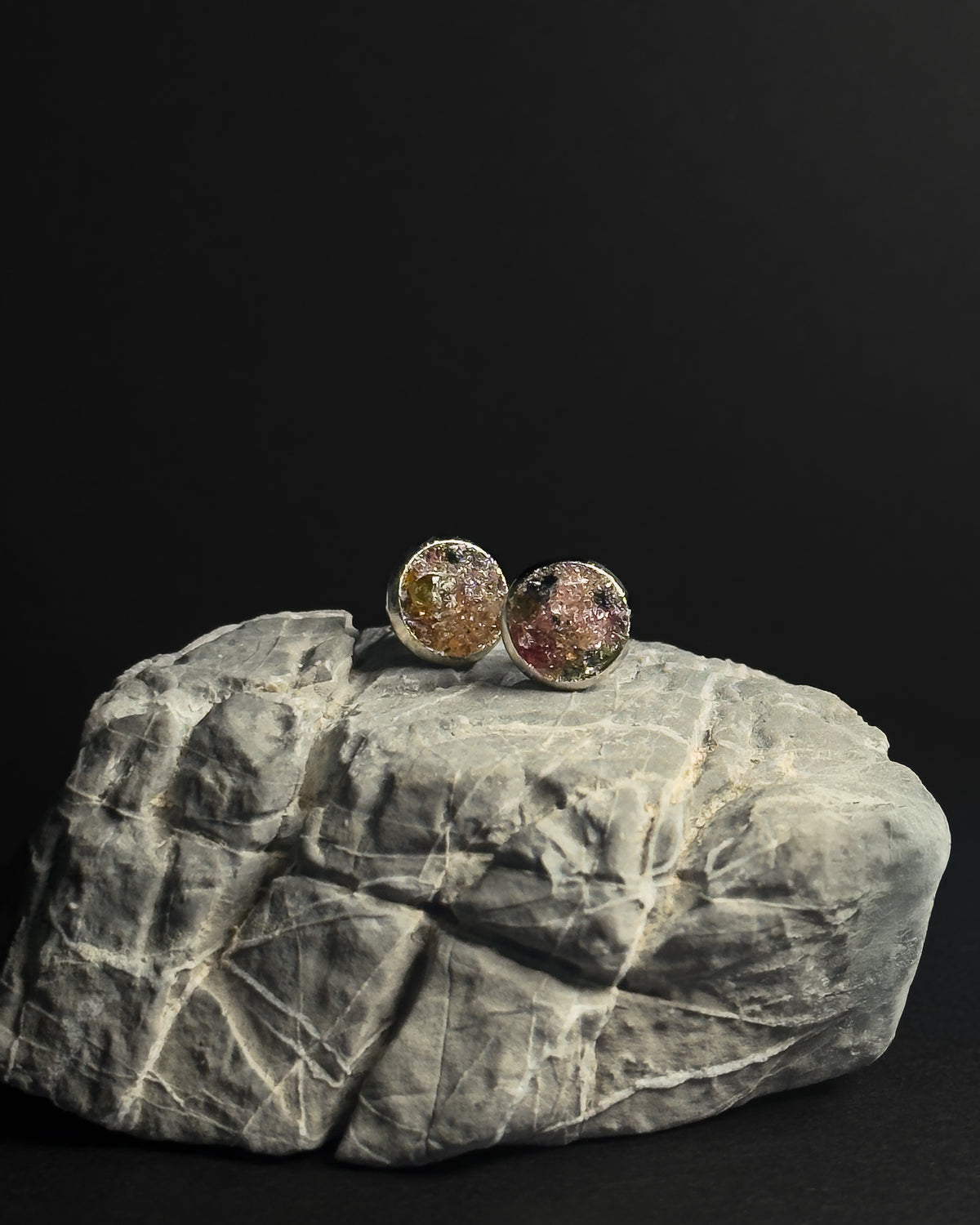 Two round stud earrings with textured, crushed rainbow tourmaline inlay displayed on a gray rock against a solid black background.