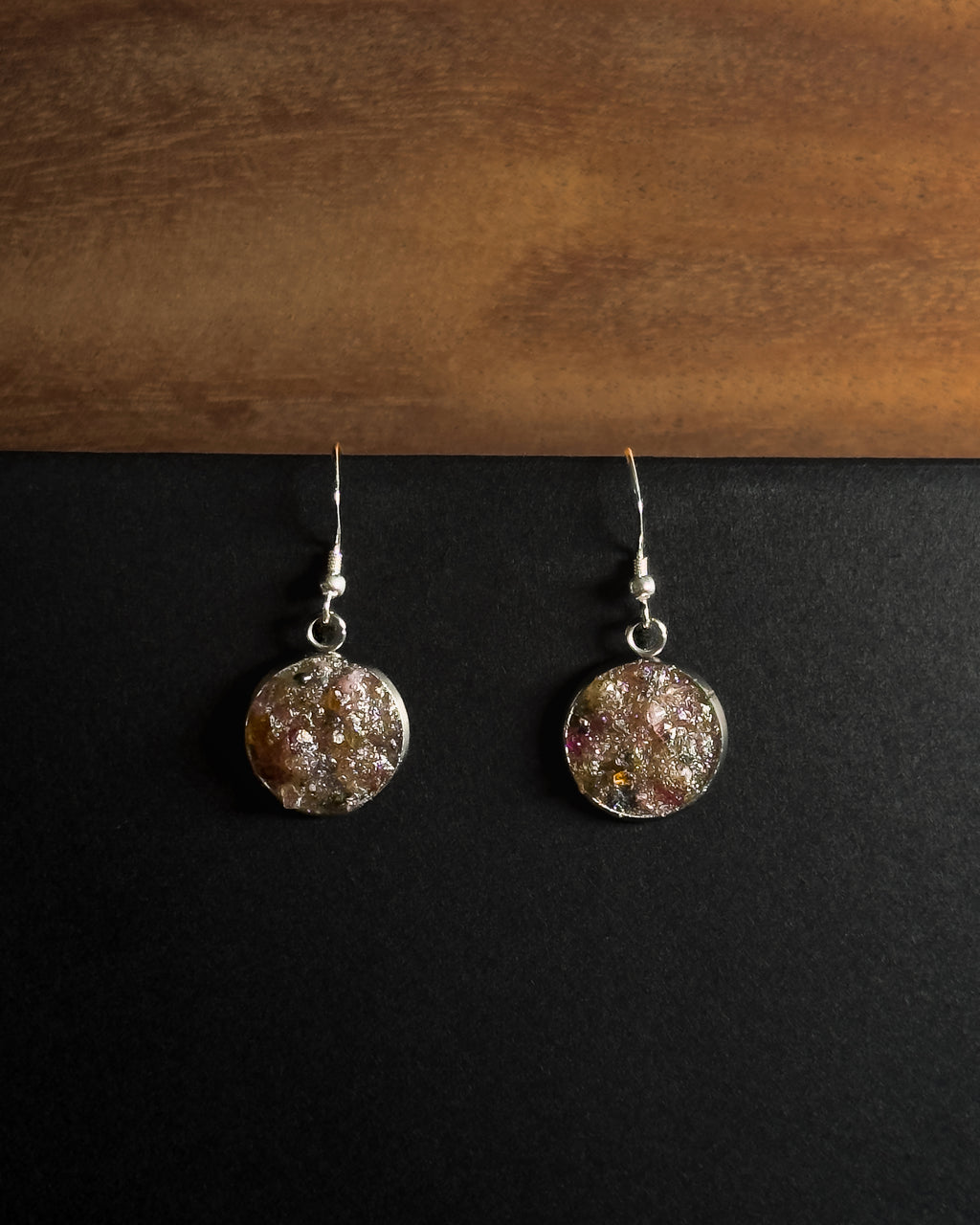 A pair of silver-toned dangle earrings with round, textured, multicolored crushed rainbow tourmaline inlay pendants resting on a black surface below a wooden board.