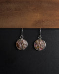 A pair of silver-toned dangle earrings with round, textured, multicolored crushed rainbow tourmaline inlay pendants resting on a black surface below a wooden board.