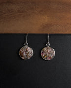 A pair of silver-toned dangle earrings with round, textured, multicolored crushed rainbow tourmaline inlay pendants resting on a black surface below a wooden board.