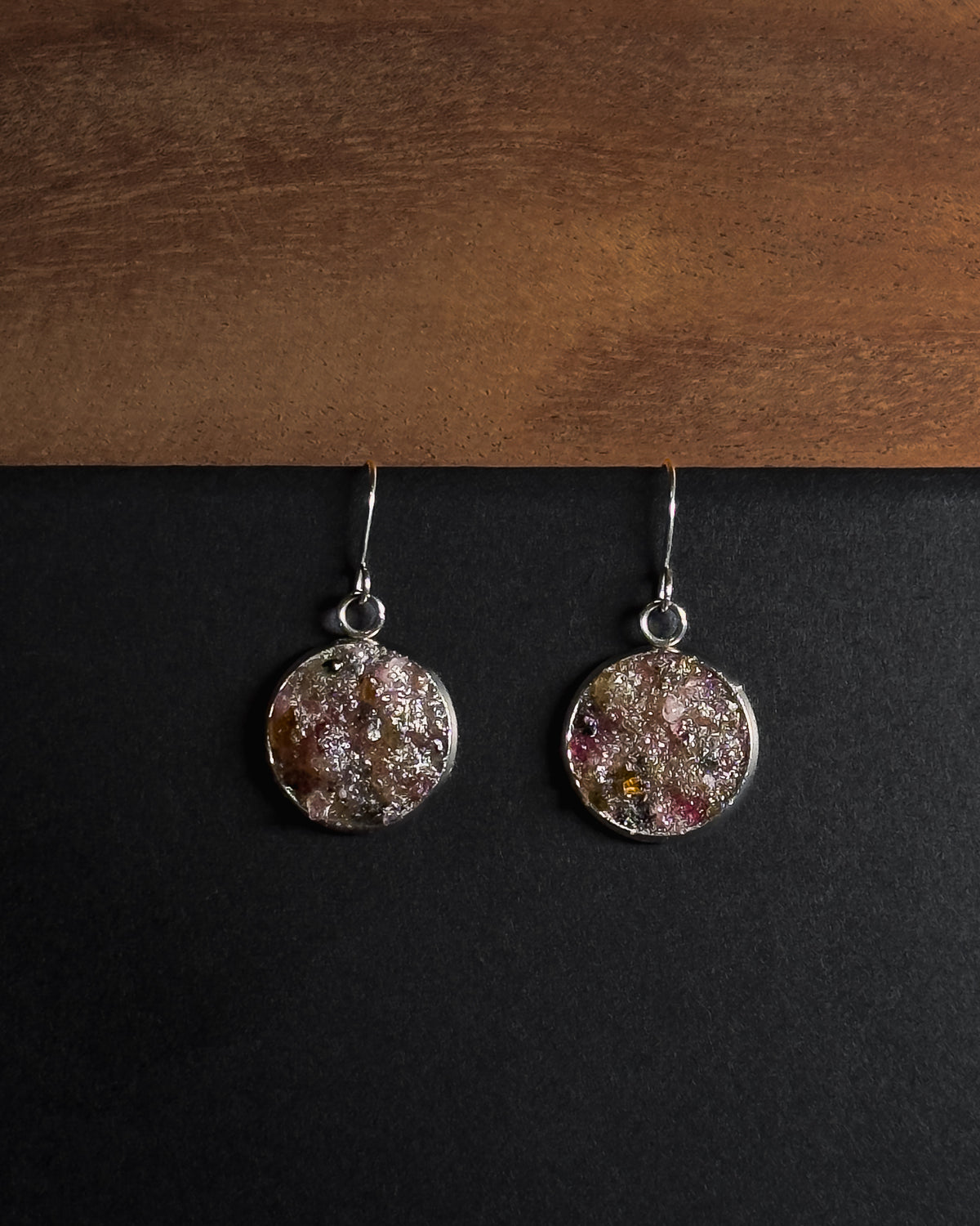 A pair of silver-toned dangle earrings with round, textured, multicolored crushed rainbow tourmaline inlay pendants resting on a black surface below a wooden board.