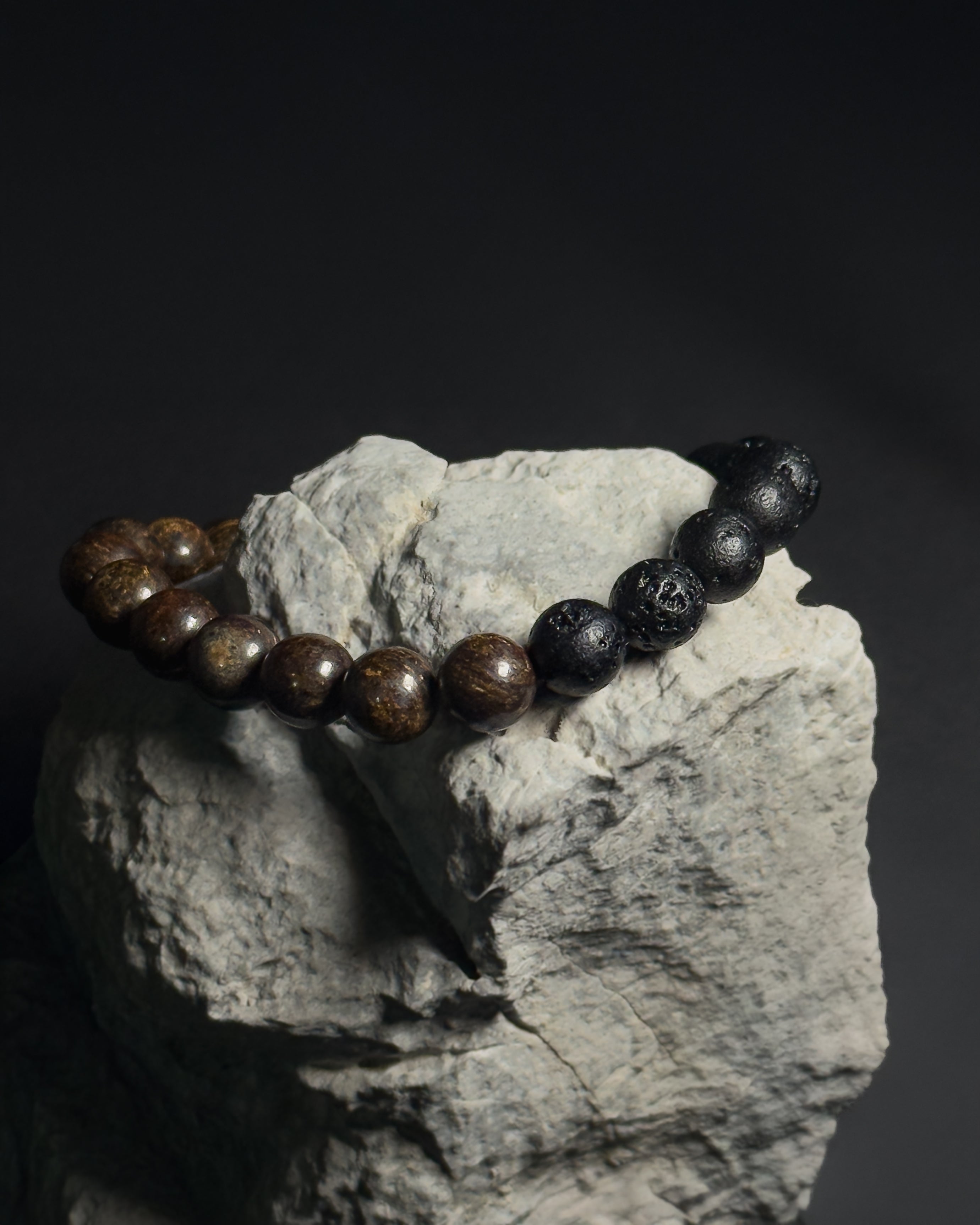 Handmade Trailborn Bracelet featuring 8mm bronzite and lava stone beads with adjustable sliding knot closure on grey rock and dark background.