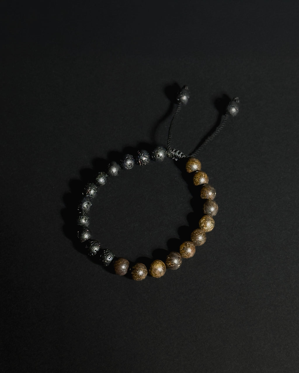 A beaded bracelet featuring natural  lava stone and bronzite , with a black adjustable cord closure, displayed on a dark background.