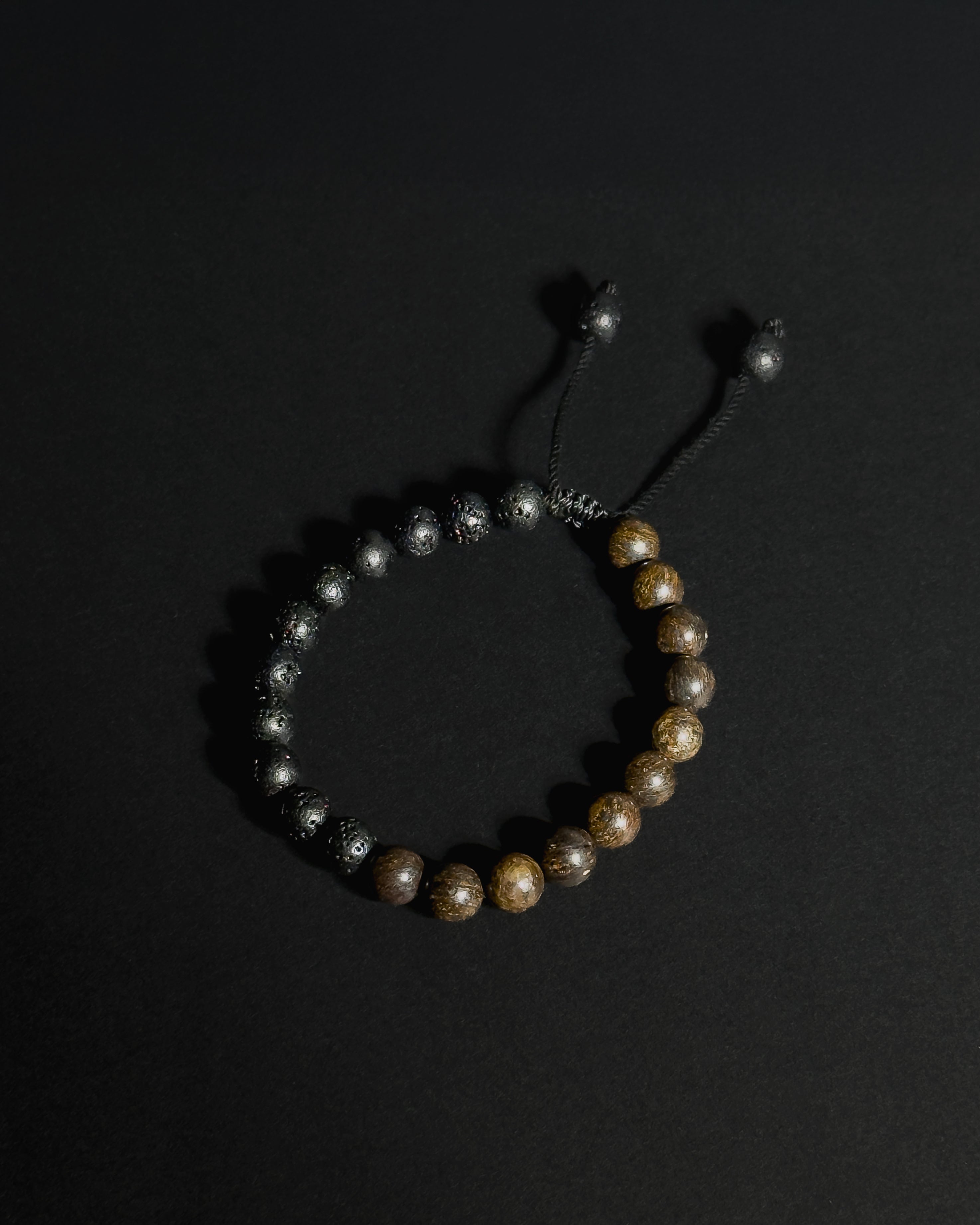 A beaded bracelet featuring natural  lava stone and bronzite , with a black adjustable cord closure, displayed on a dark background.