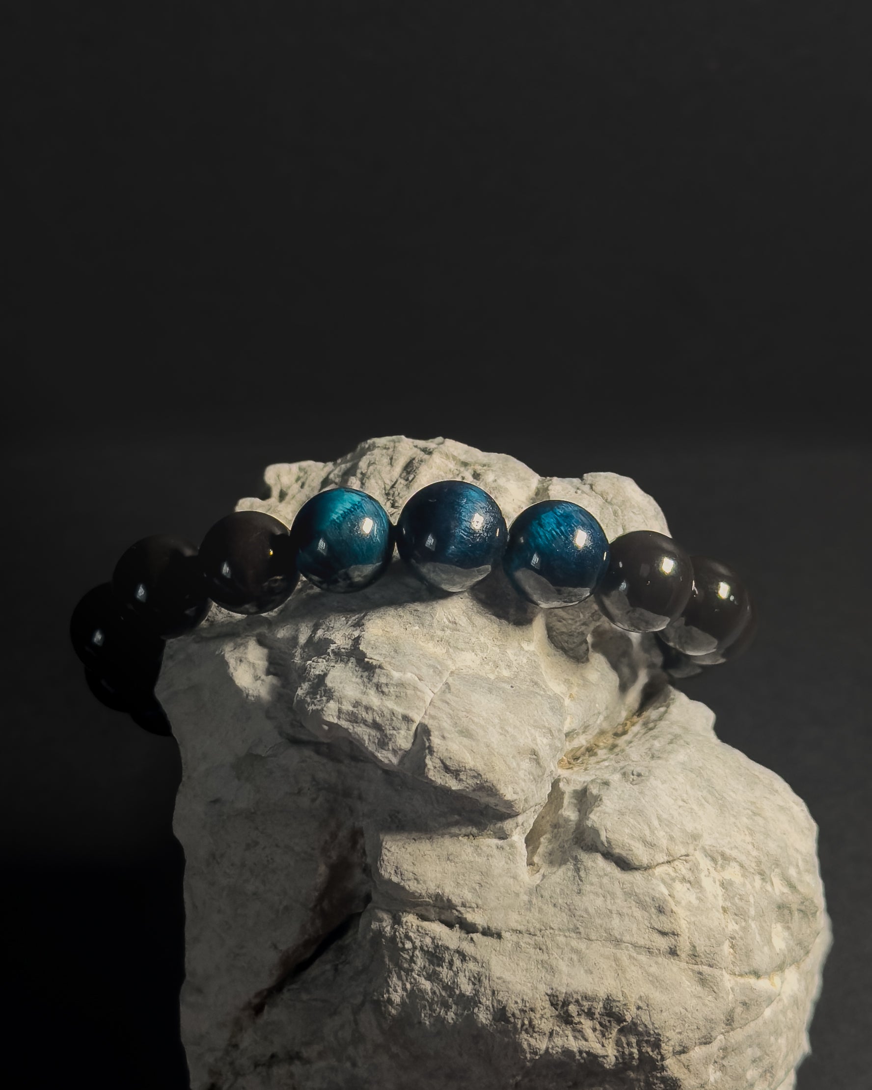 Bracelet with blue and black beads on a rock against a dark background. Black beaded bracelet with blue tiger eye.