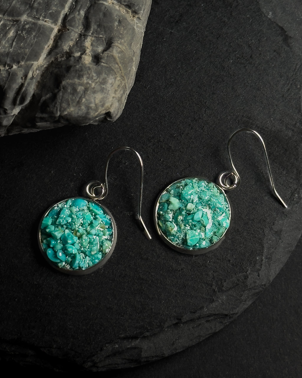 A pair of round drop earrings with textured crushed turquoise inlays set in silver-tone metal, hanging from silver fishhook wires on a dark slate surface.