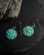 A pair of round drop earrings with textured crushed turquoise inlays set in silver-tone metal, hanging from silver fishhook wires on a dark slate surface.
