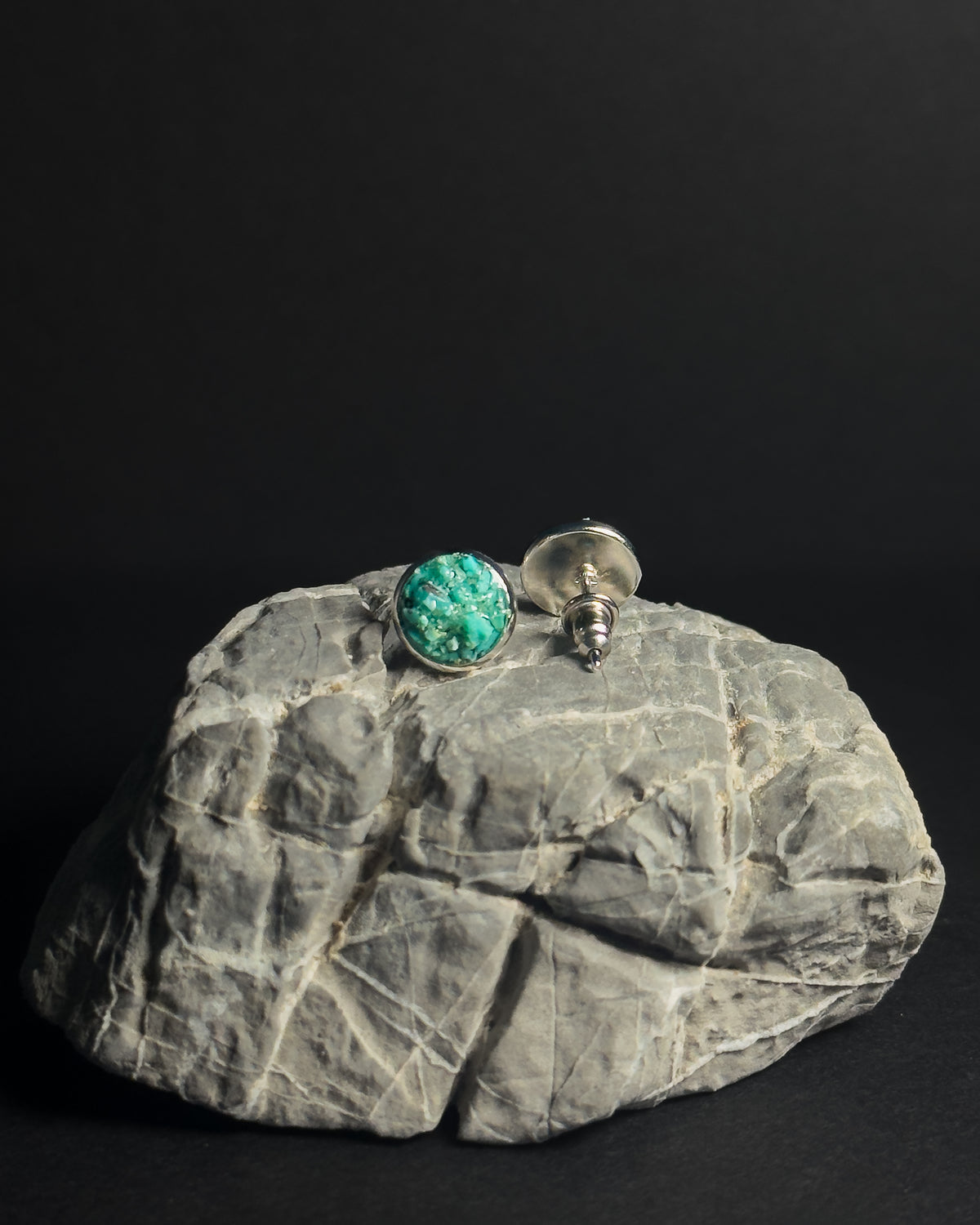 A pair of silver stud earrings, one featuring a round crushed turquoise stone inlay and the other showing the silver backing, displayed on a textured gray rock against a solid black background.