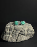 A pair of round stud earrings with vibrant crushed turquoise stone inlay  in silver bezel settings, displayed on a textured gray rock against a solid black background.