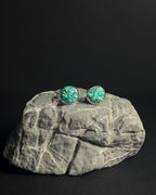 A pair of round stud earrings with vibrant crushed turquoise stone inlay  in silver bezel settings, displayed on a textured gray rock against a solid black background.