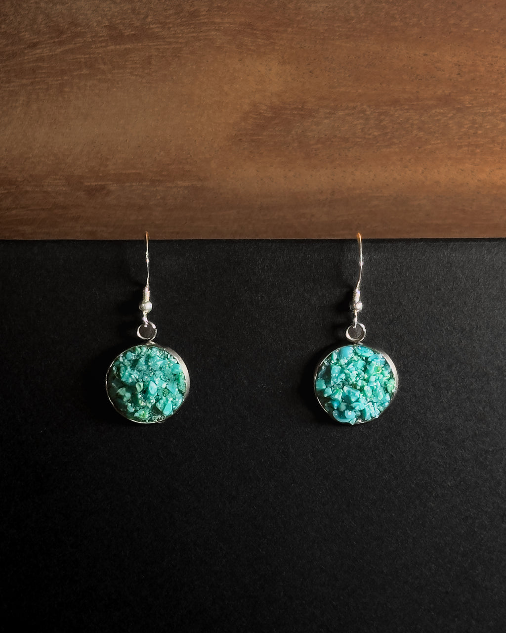 A pair of silver dangle earrings with round crushed turquoise  stone pendants, displayed on a split black and brown wooden background.