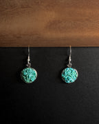 A pair of silver dangle earrings with round crushed turquoise  stone pendants, displayed on a split black and brown wooden background.