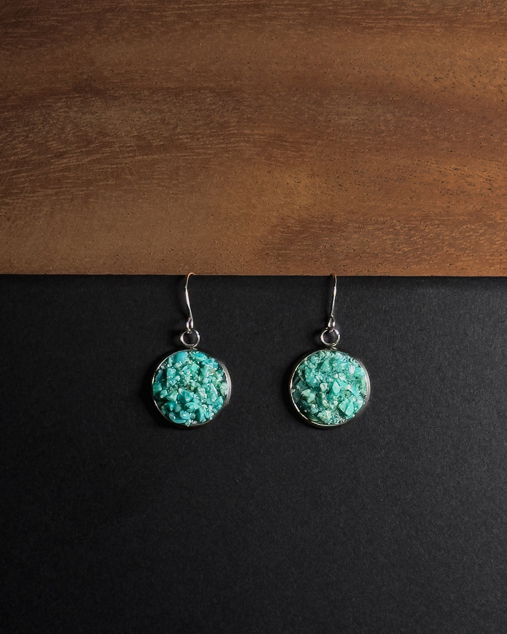 A pair of silver dangle earrings with round crushed turquoise  stone pendants, displayed on a split black and brown wooden background.