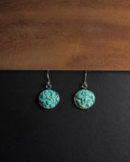 A pair of silver dangle earrings with round crushed turquoise  stone pendants, displayed on a split black and brown wooden background.