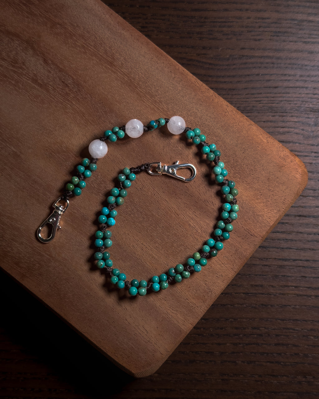 A beaded phone charm keychain with turquoise-blue and white beads, with silver toned stainless steel clasp displayed on a wooden board over a dark wood surface.