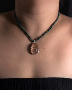 A close-up of a person's neck and chest wearing a beaded necklace with a teardrop-shaped brown gemstone pendant.