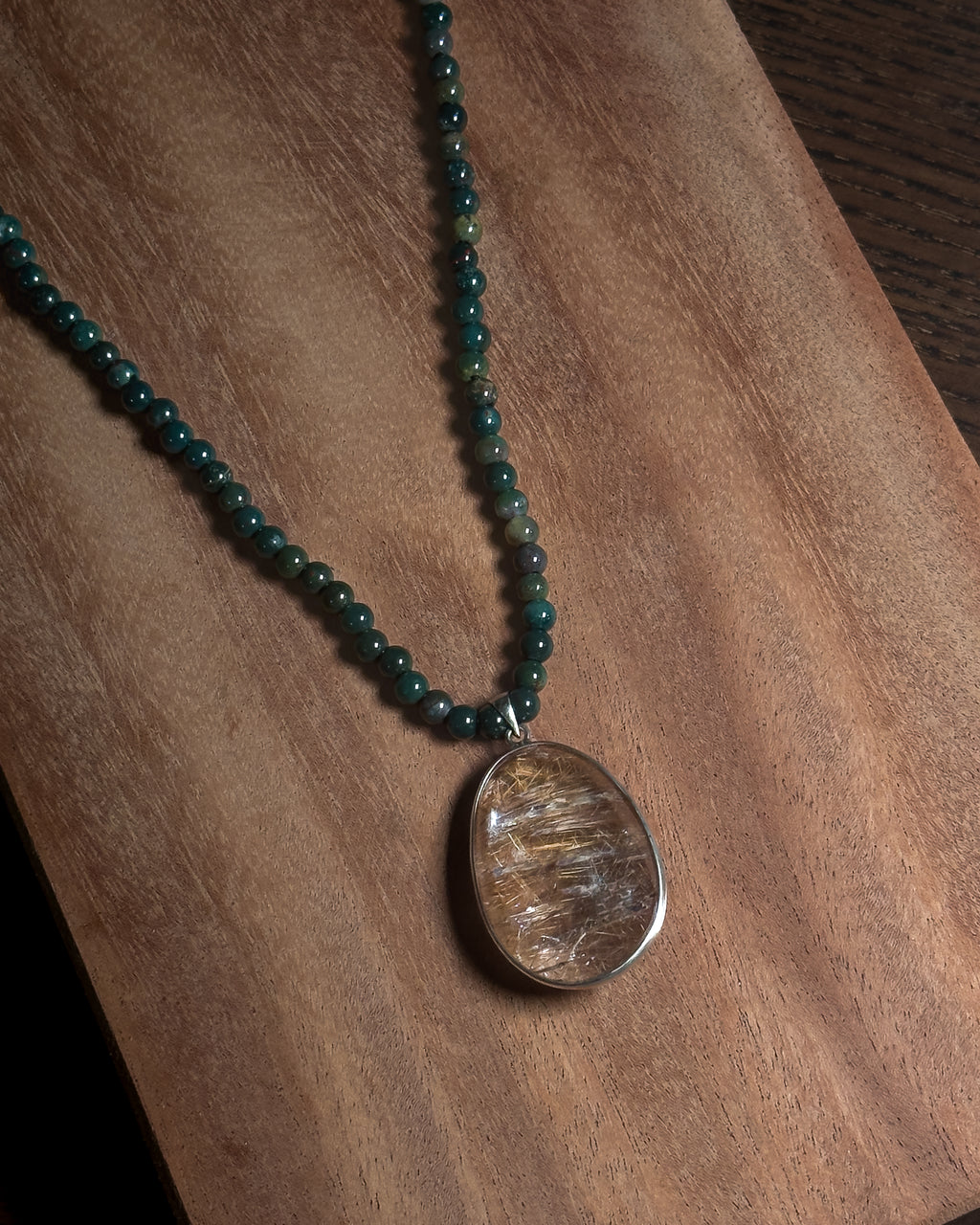 A beaded necklace with dark green beads and a large oval rutilated quartz pendant with gold-toned swirls, displayed on a wooden surface.