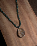 A beaded necklace with dark green beads and a large oval rutilated quartz pendant with gold-toned swirls, displayed on a wooden surface.