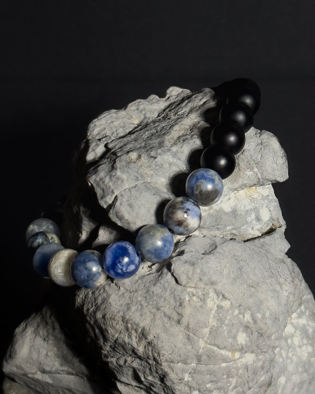 A beaded bracelet with black matte onyx agate beads and sodalite stone beads, draped over a gray rock against a dark background.