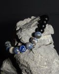 A beaded bracelet with black matte onyx agate beads and sodalite stone beads, draped over a gray rock against a dark background.