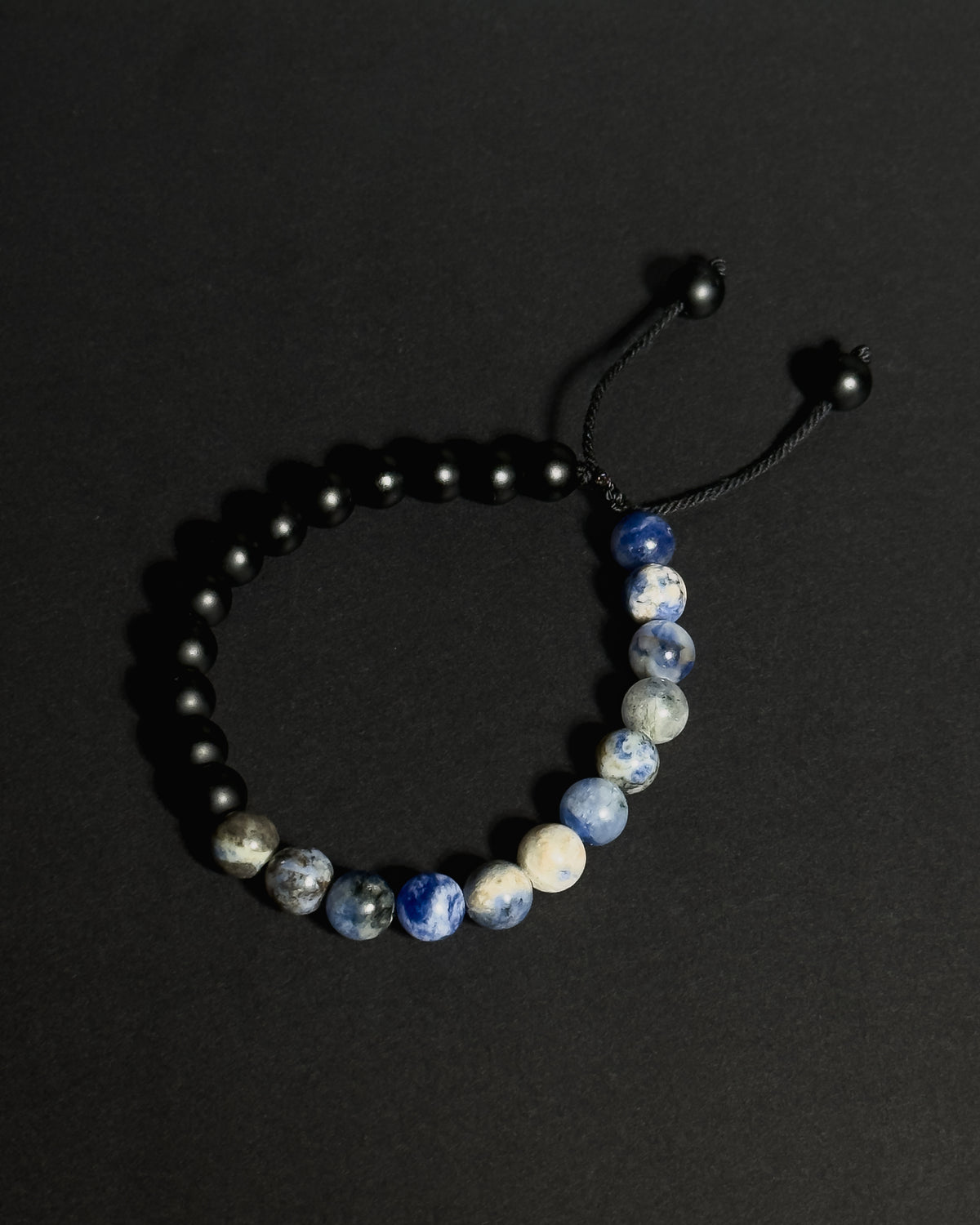 A beaded bracelet with matte black beads on one side and a section of blue, white, and gray sodalite beads on the other, connected by a black adjustable cord with black beads.