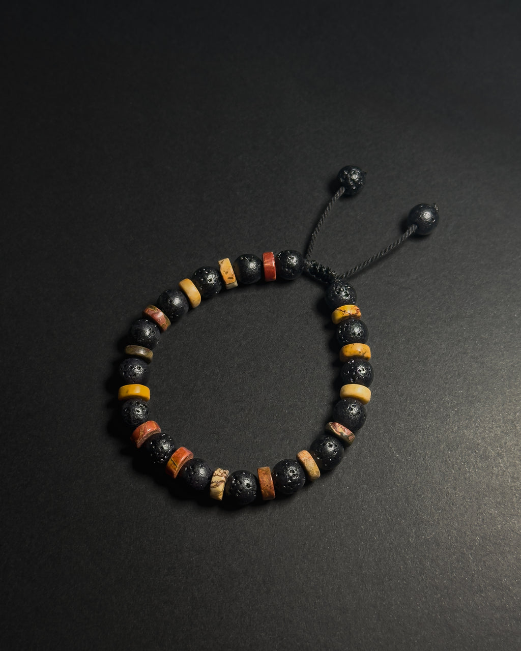 A beaded bracelet with black lava stones, yellow and brown picasso jasper accent beads, and a black adjustable cord closure, displayed on a dark background.