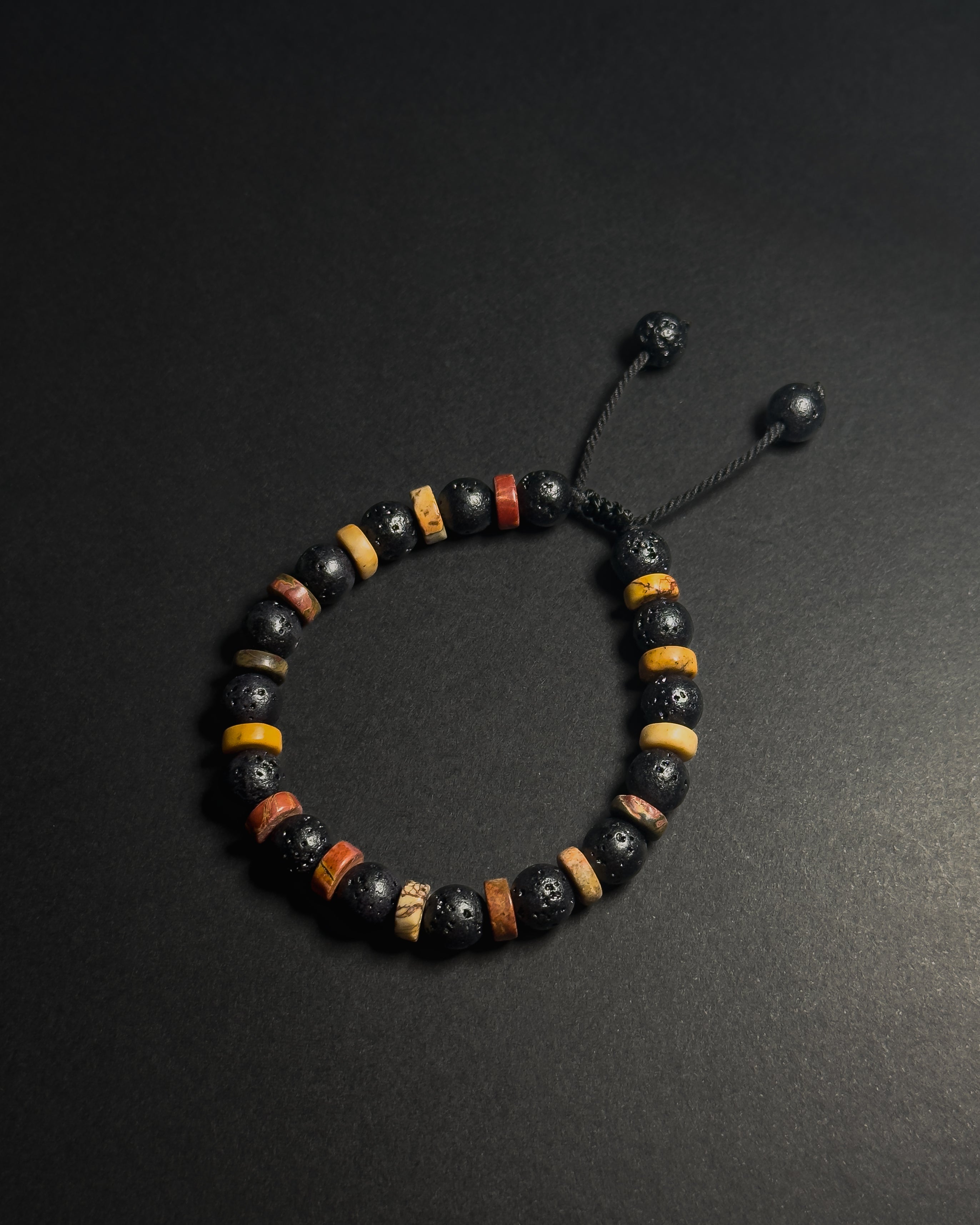 A beaded bracelet with black lava stones, yellow and brown picasso jasper accent beads, and a black adjustable cord closure, displayed on a dark background.